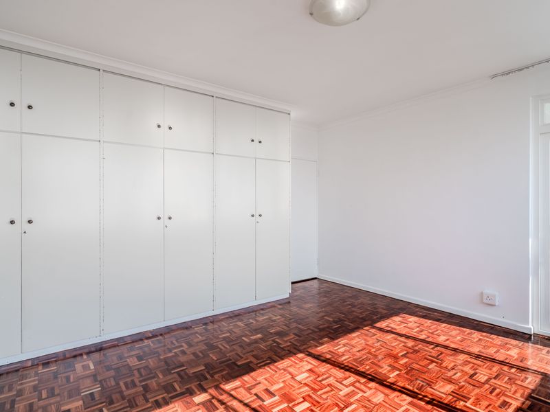 A bright, empty bedroom featuring classic parquet flooring, extensive built-in cupboard storage, and a glass-panelled door leading to an outdoor area.
