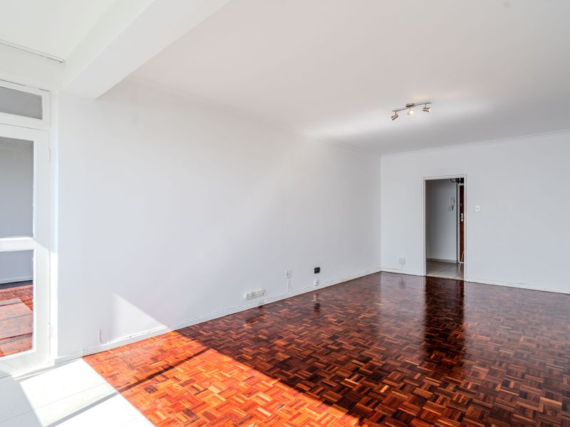 A bright, empty living room featuring polished parquet flooring, neutral white walls, and a glass-paneled door leading to an outdoor area.