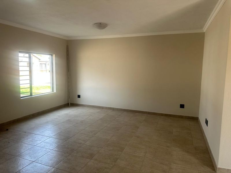 A spacious, empty living area featuring neutral tiled flooring, clean walls, and a window with security bars, offering a blank canvas for personalization.