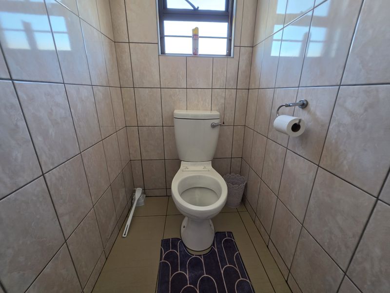 A compact bathroom featuring full-height wall tiling, a standard toilet, and a window with security bars.