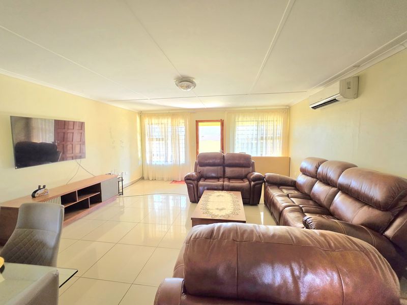 A spacious living room featuring tiled flooring, a wall-mounted air conditioning unit, and natural light through large windows.
