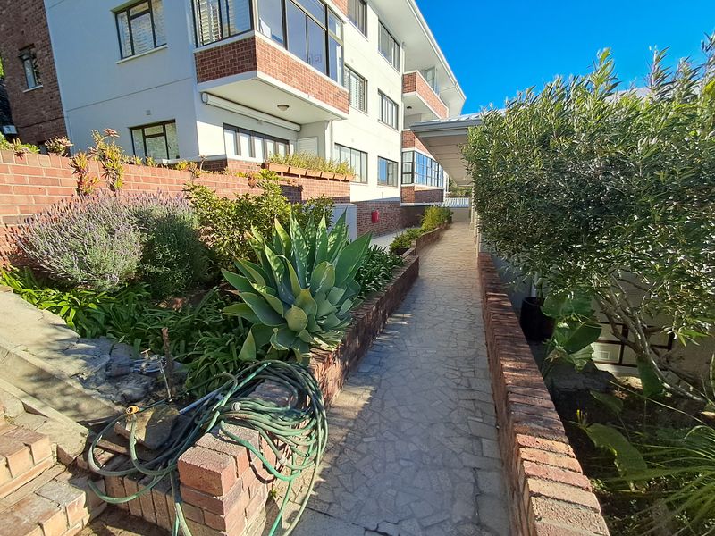 A well-maintained exterior walkway featuring brick retaining walls, stone paving, and established garden beds alongside the apartment building facade.