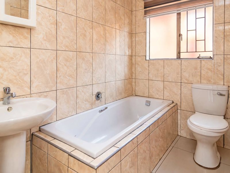 A functional bathroom featuring a built-in bathtub, pedestal washbasin, and toilet, finished with neutral marble-effect wall tiles and a security window grille.