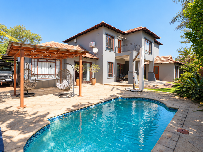 A well-maintained rear exterior featuring a swimming pool, paved patio area with a wooden pergola, and a two-story house with a balcony.