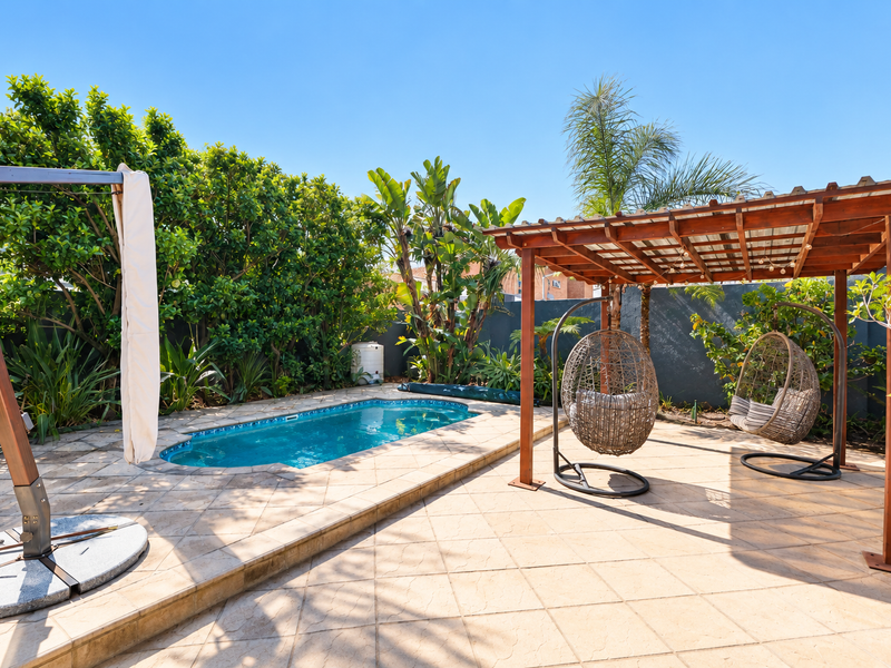A well-maintained outdoor pool area featuring a paved patio, a timber pergola, and established garden landscaping.