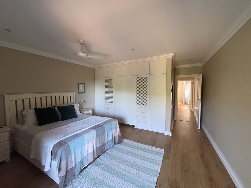 Spacious bedroom featuring light laminate flooring, extensive built-in cabinetry with integrated storage, and a ceiling fan, leading into a bright hallway.