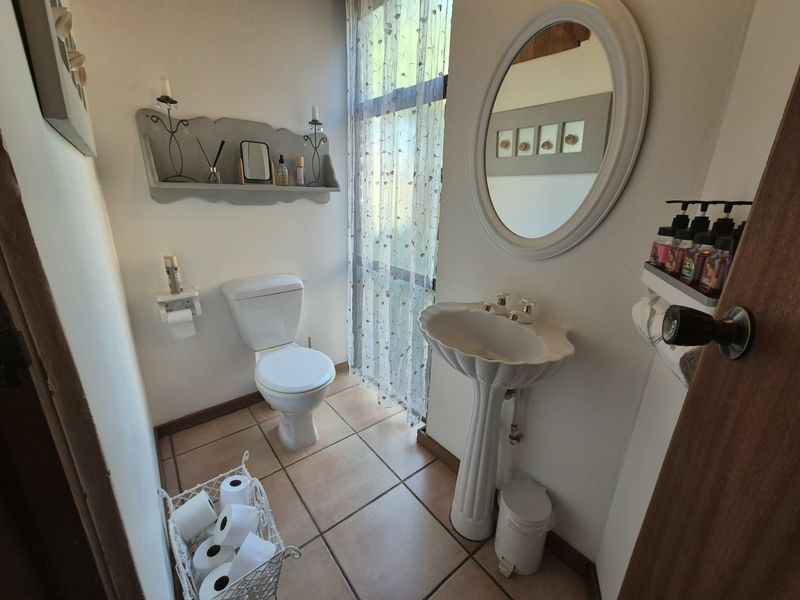 A compact bathroom featuring a pedestal basin, toilet, and tiled flooring, illuminated by a large window with sheer curtains.