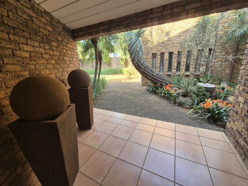 A covered patio area featuring brick walls and tiled flooring, overlooking a landscaped garden with a paved walkway.