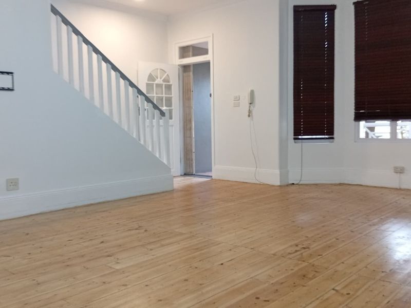 A bright, vacant living room featuring polished timber flooring, a staircase with a white balustrade, and a bay window with fitted venetian blinds.