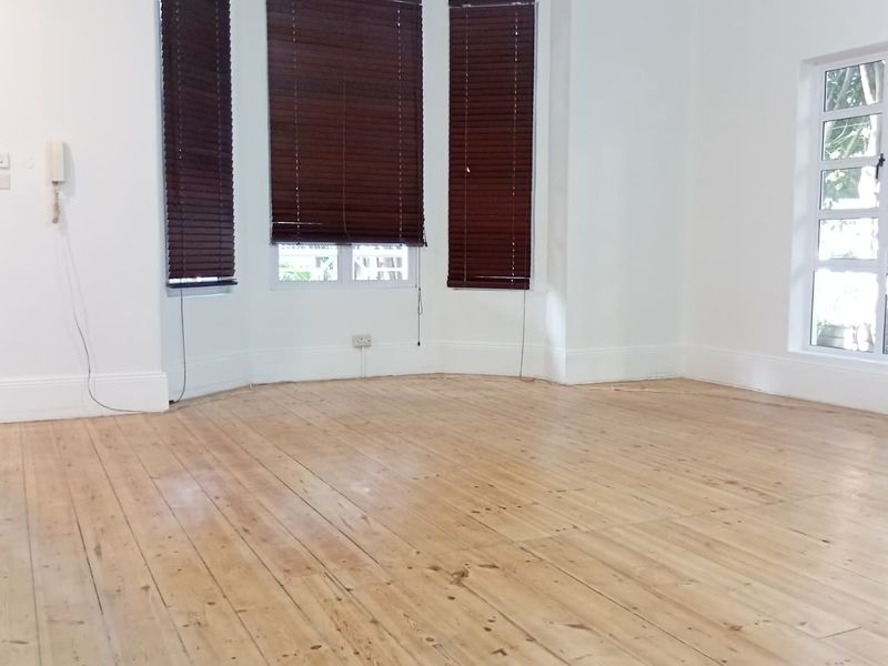 A bright, empty living room featuring polished timber flooring, a large bay window with venetian blinds, and classic cornicing details.