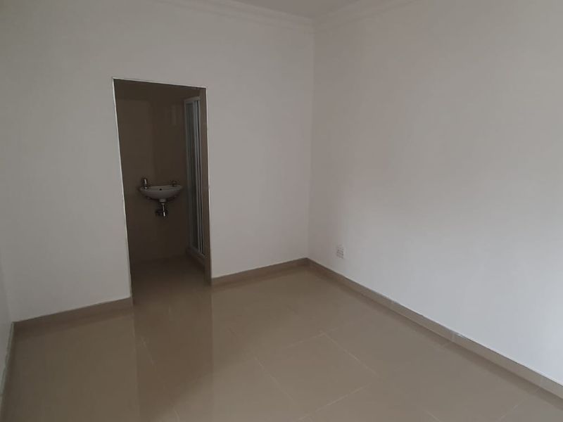A clean, empty bedroom featuring neutral tiled flooring, white walls, and direct access to an en-suite bathroom.