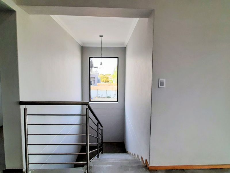 A bright, minimalist hallway featuring a staircase with a modern stainless steel balustrade and a large window providing natural light.