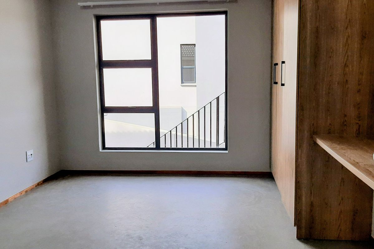 A clean, empty bedroom featuring built-in wooden cupboards, a built-in desk, and a large window providing natural light.