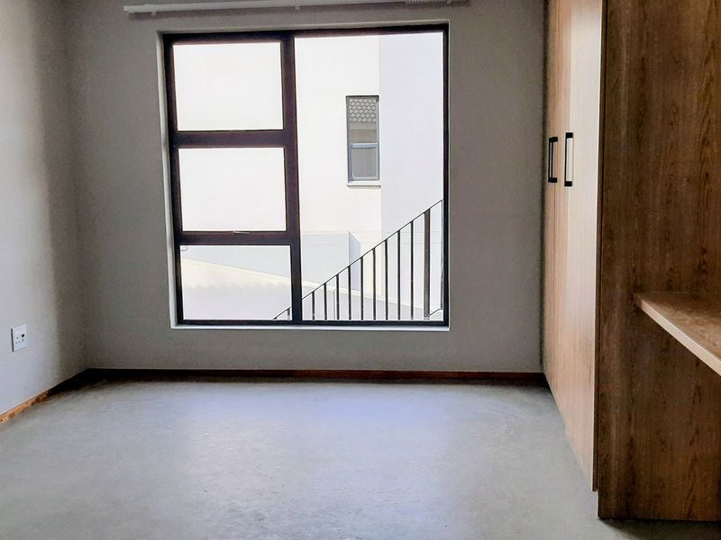 A clean, empty bedroom featuring built-in wooden cupboards, a built-in desk, and a large window providing natural light.