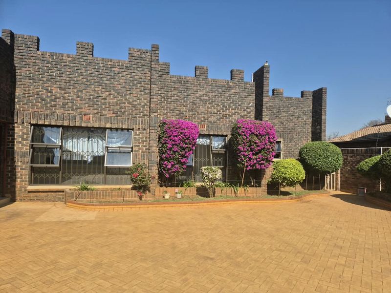 Front elevation of a face brick house featuring a distinctive parapet wall design, paved driveway, and established garden beds with security window bars.