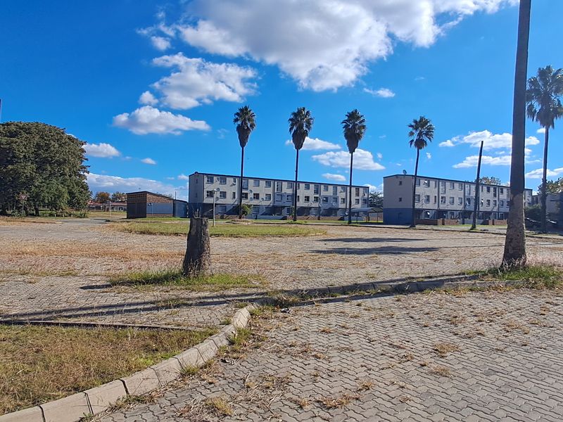 Exterior view of a multi-story apartment complex with a large paved parking area and mature palm trees.