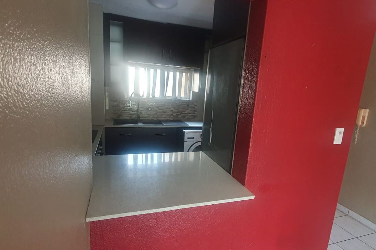 A compact kitchen featuring dark cabinetry, a stone countertop, and a tiled backsplash, viewed through a service opening from the adjacent living area.