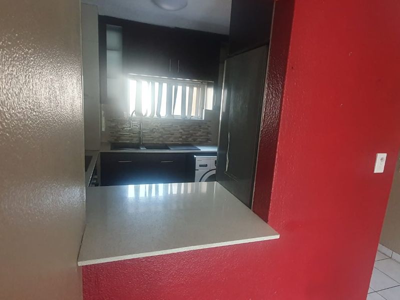 A compact kitchen featuring dark cabinetry, a stone countertop, and a tiled backsplash, viewed through a service opening from the adjacent living area.