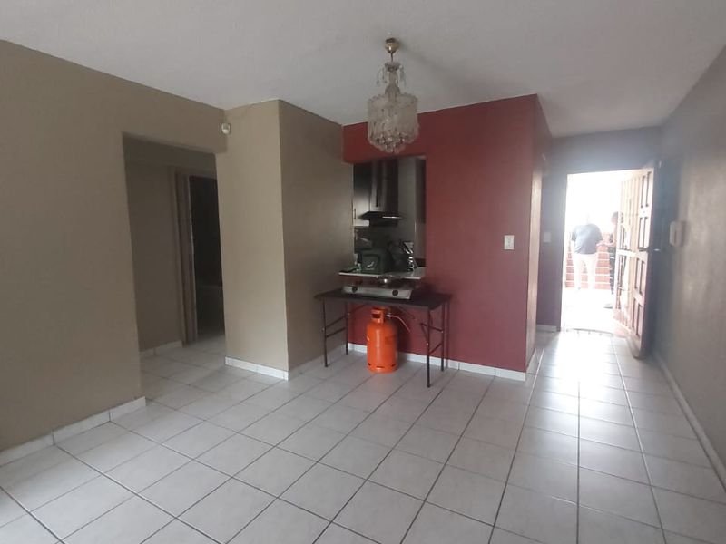 An open-plan living area featuring tiled flooring and a neutral color palette, leading to an exterior entrance and a small kitchen alcove.