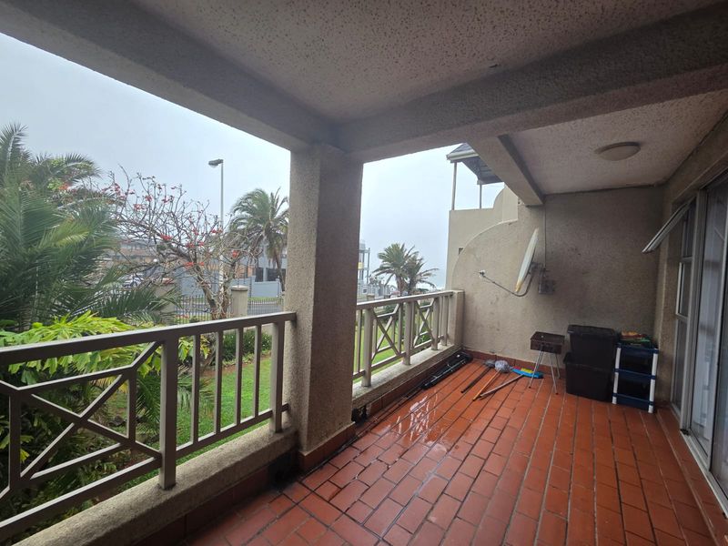 A covered balcony featuring terracotta tile flooring, concrete structural pillars, and a safety railing, overlooking the surrounding garden area.