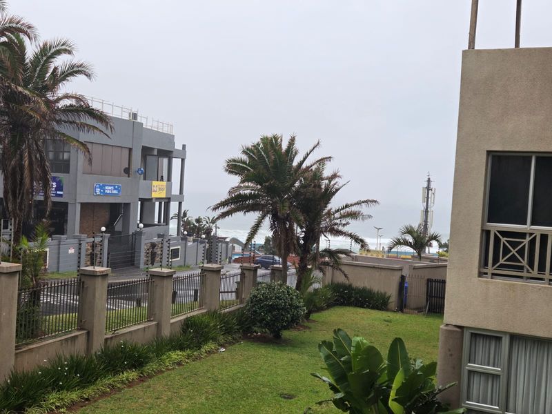An elevated view overlooking a landscaped garden, perimeter fencing, and the surrounding neighborhood with a glimpse of the ocean in the distance.