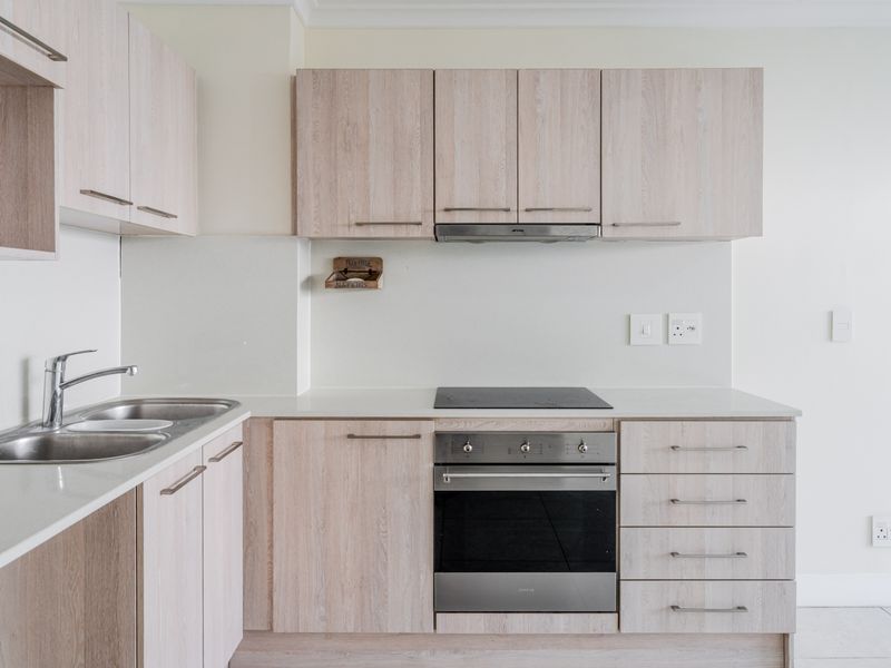 A clean, modern kitchen featuring light wood-grain cabinetry, stone countertops, a built-in oven, electric hob, and stainless steel sink.
