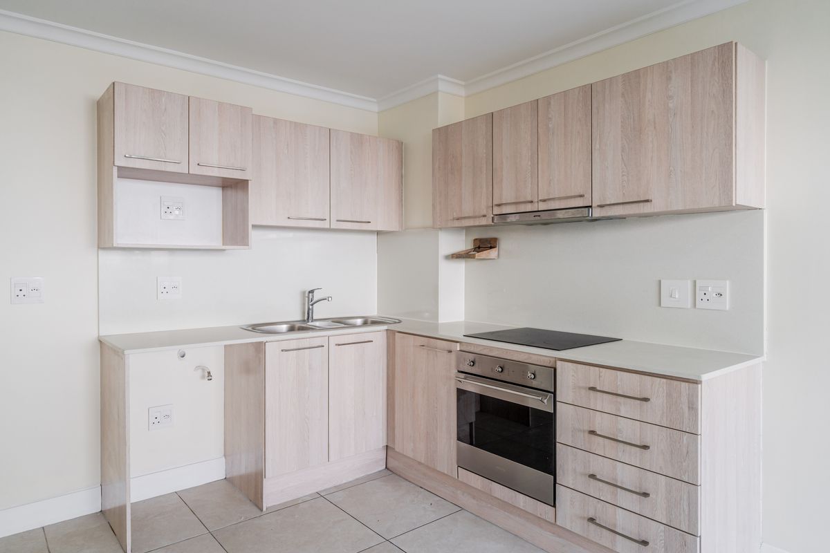 A clean, modern kitchen featuring light wood-grain cabinetry, integrated oven and hob, stainless steel sink, and neutral tiled flooring.