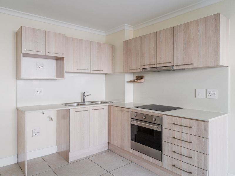 A clean, modern kitchen featuring light wood-grain cabinetry, integrated oven and hob, stainless steel sink, and neutral tiled flooring.