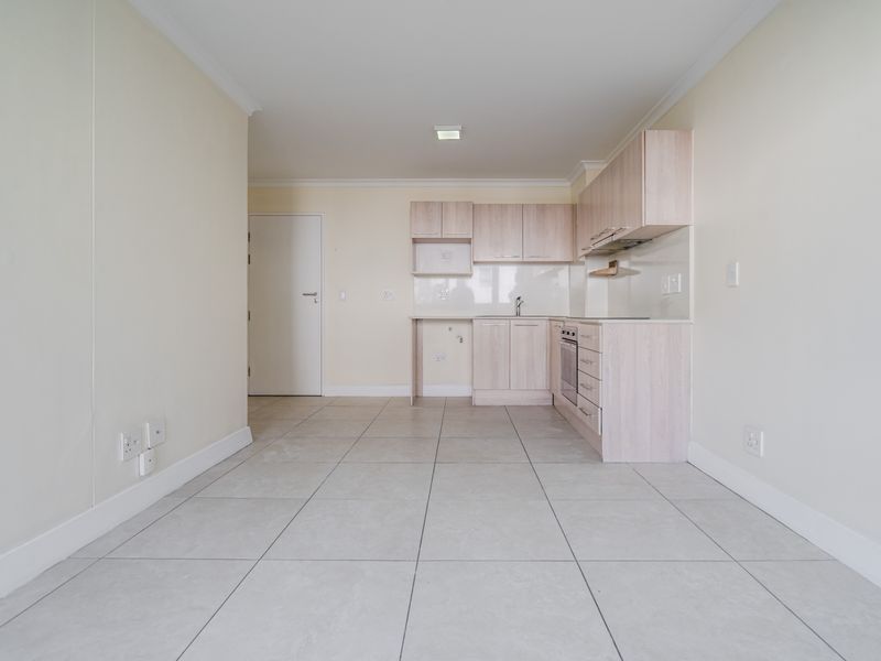 An open-plan living and kitchen area featuring light-toned built-in cabinetry, tiled flooring, and integrated kitchen appliances in a clean, vacant space.