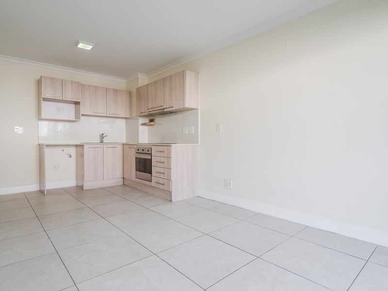 An open-plan kitchen area featuring light wood-finish cabinetry, integrated appliances, and neutral tiled flooring in a clean, well-maintained space.