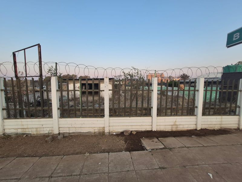A vacant residential plot secured by a concrete perimeter wall, steel palisade fencing, and razor wire, featuring a paved sidewalk frontage.