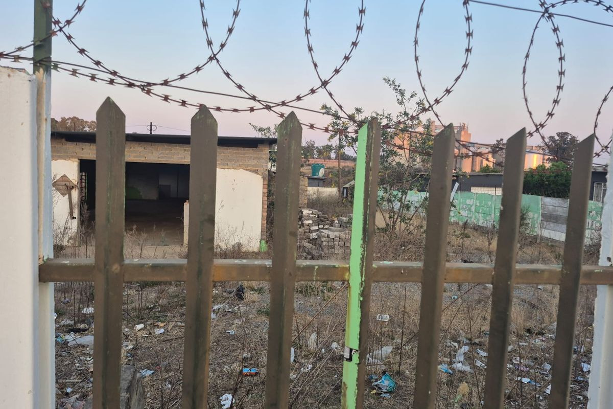 A view of a vacant residential plot featuring a metal picket fence with razor wire and an unfinished concrete block structure in the background.