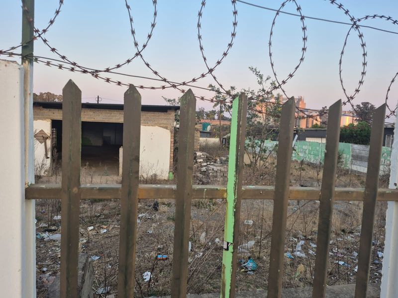 A view of a vacant residential plot featuring a metal picket fence with razor wire and an unfinished concrete block structure in the background.