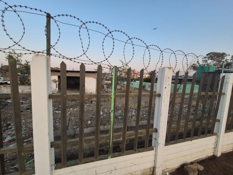 View of a vacant residential plot secured with a metal palisade fence and razor wire, showing signs of neglect and accumulated debris.