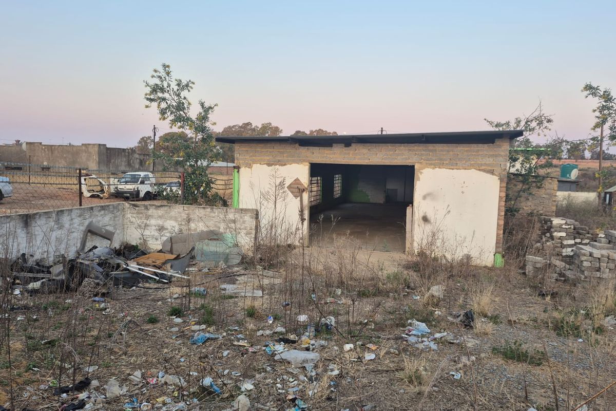A view of a vacant residential plot featuring a small, unfinished brick structure and significant debris on the property grounds.