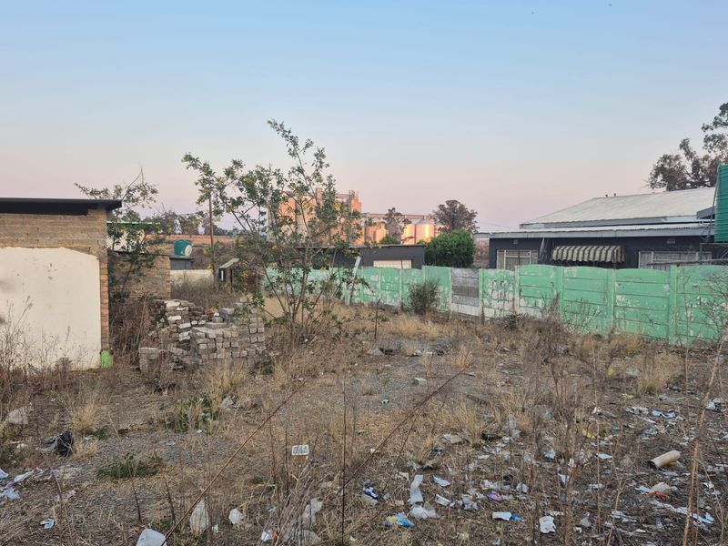A vacant residential plot featuring a precast concrete boundary wall, water tank, and significant overgrown vegetation with accumulated debris.