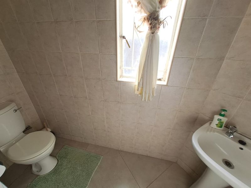 A functional bathroom featuring neutral ceramic wall and floor tiles, a standard pedestal basin, and a toilet with a window for natural light.