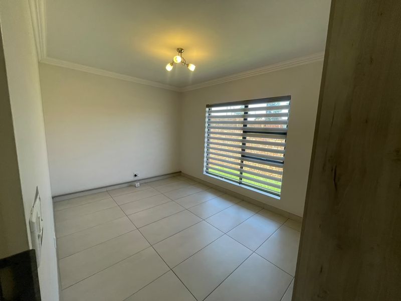 A clean, empty bedroom featuring neutral tiled flooring, modern ceiling lighting, and a window fitted with zebra blinds.