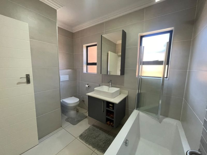 Modern bathroom featuring a wall-mounted toilet, vanity with vessel sink, and a bathtub with a glass shower screen, finished with neutral wall tiles.