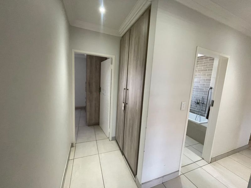 A clean, modern hallway featuring neutral tiled flooring, built-in wood-finish cupboards, and recessed ceiling lighting, providing access to adjacent rooms.
