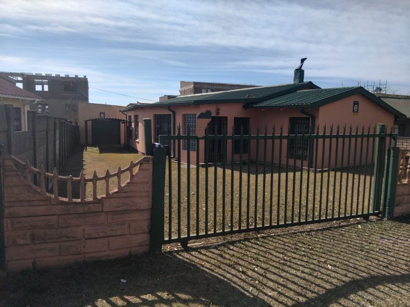 Front view of a single-story house featuring a metal security gate, precast boundary wall, and a pitched metal roof with a chimney.