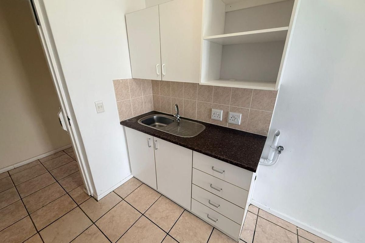 A compact kitchen area featuring white cabinetry, dark laminate countertops, a stainless steel sink, and neutral ceramic floor tiles.
