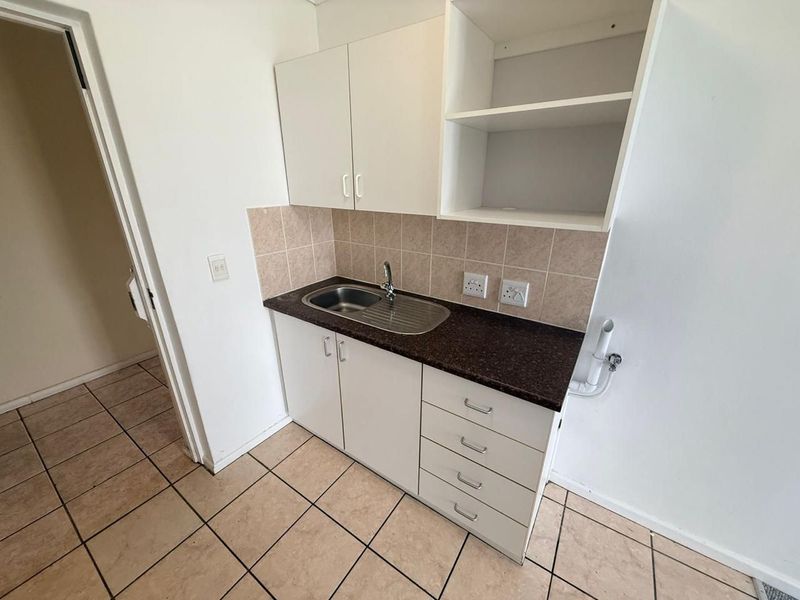 A compact kitchen area featuring white cabinetry, dark laminate countertops, a stainless steel sink, and neutral ceramic floor tiles.