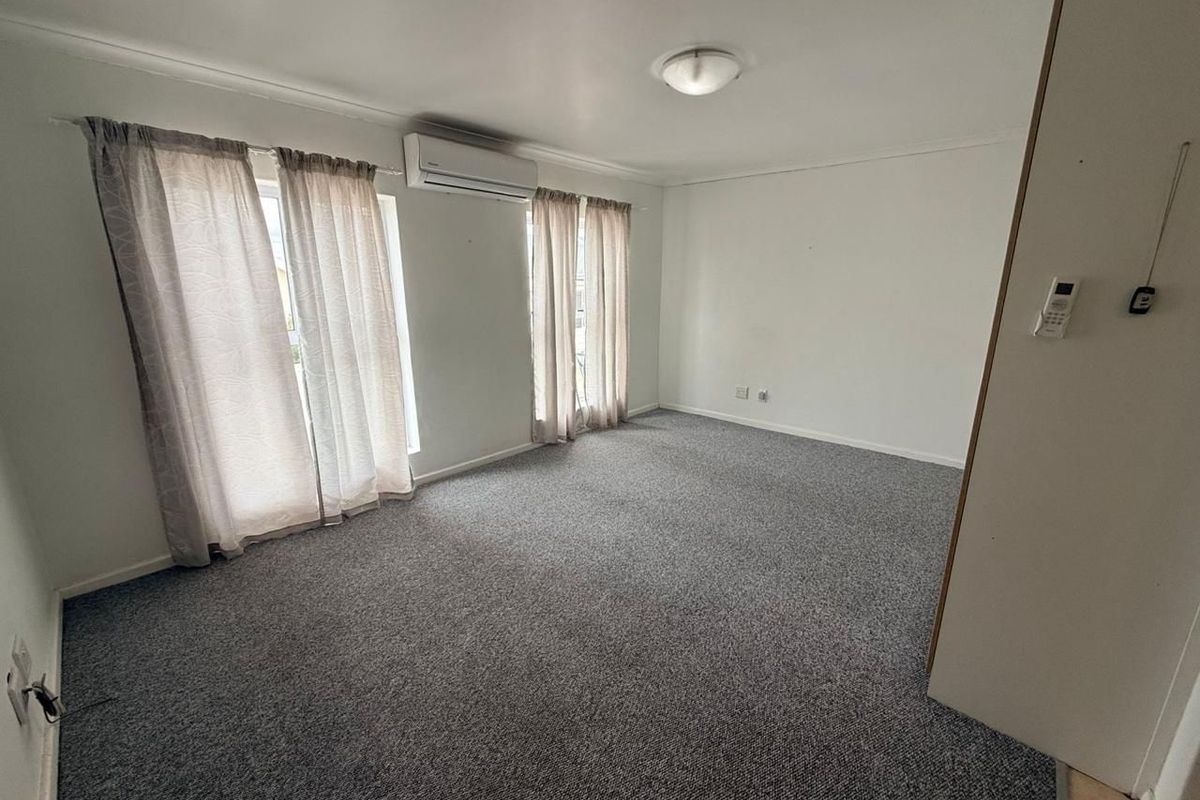 An empty living room featuring grey carpet flooring, a wall-mounted split-system air conditioner, and natural light through large windows.
