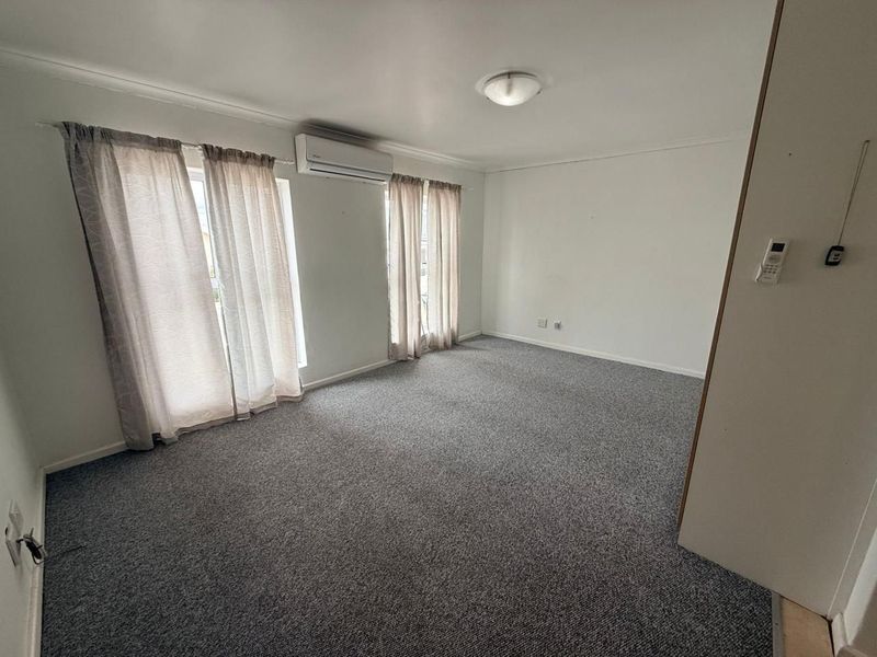 An empty living room featuring grey carpet flooring, a wall-mounted split-system air conditioner, and natural light through large windows.