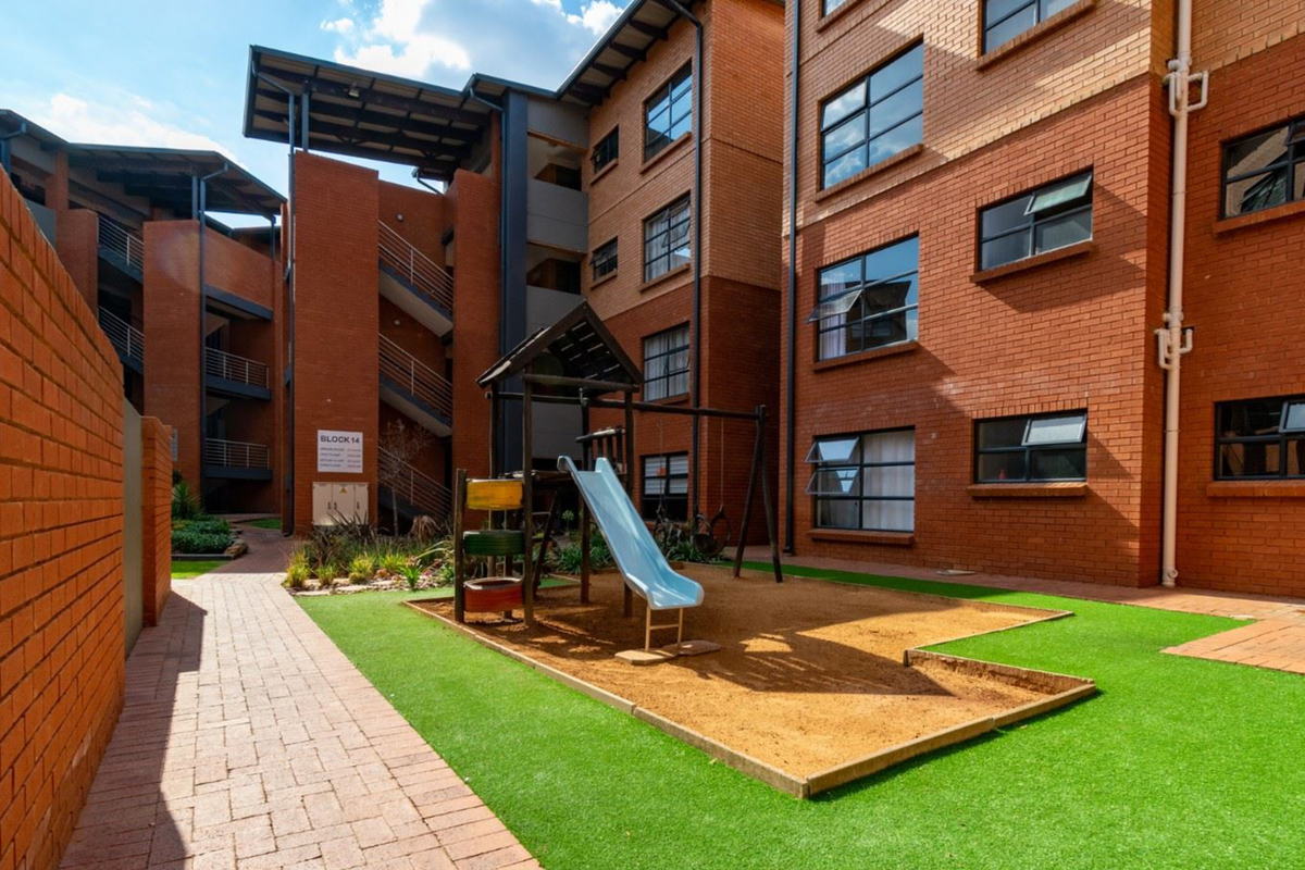A well-maintained communal courtyard area featuring face brick townhouse facades, a paved walkway, artificial grass, and a children's play area with a sand pit.