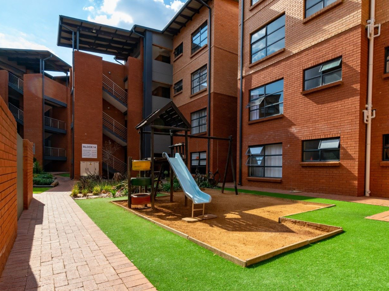 A well-maintained communal courtyard area featuring face brick townhouse facades, a paved walkway, artificial grass, and a children's play area with a sand pit.