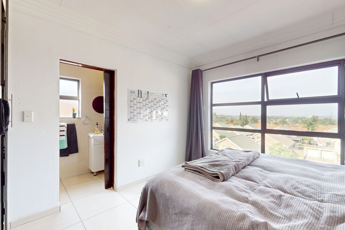 A bright bedroom featuring tiled floors, a built-in wardrobe, and an en-suite bathroom, with large windows offering an elevated view of the surrounding area.