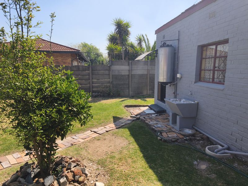 Rear exterior view of the townhouse featuring a small garden, brick pathway, external geyser, and a fixed laundry sink area.