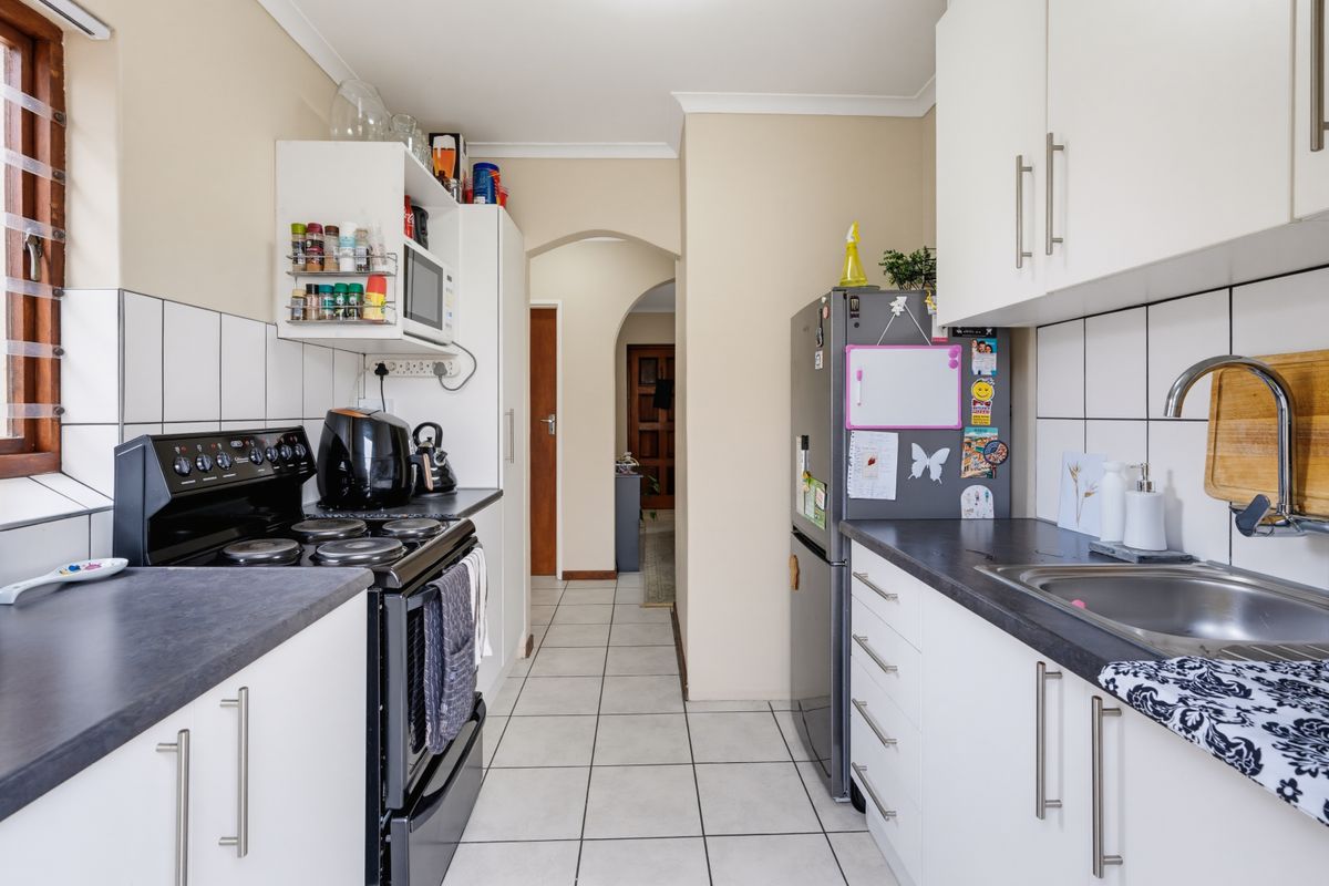 A functional galley-style kitchen featuring white cabinetry, dark laminate countertops, tiled splashbacks, and a freestanding stove, leading through an archway to the hallway.